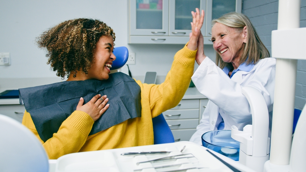 A smiling patient in a yellow sweater high-fives a cheerful dentist in a white coat inside a dental office, showing happiness and positive connection after a dental appointment. Dental tools are visible in the foreground.