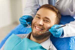 man with a beard in a dental chair smiling getting work done with a dentist behind him