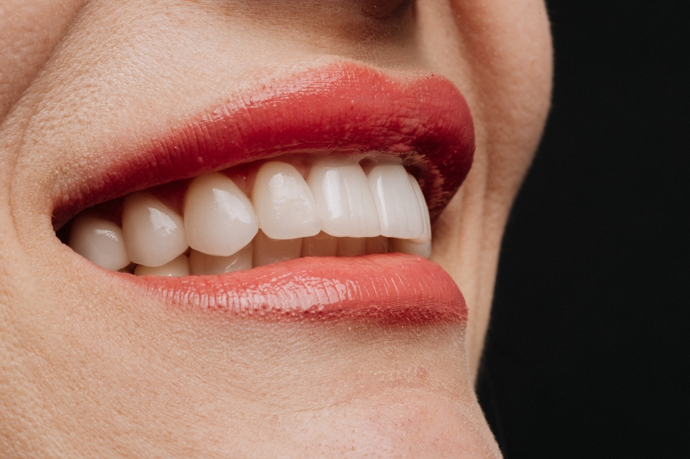 Close-up of a smiling mouth with smooth, red lipstick and straight, white teeth. The lips are slightly parted, showing the upper and lower teeth against a plain black background.