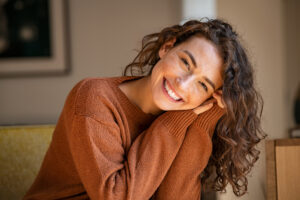 A young woman with curly brown hair smiles brightly, leaning her head on her hands. She wears a rust-colored sweater and sits indoors in soft natural light, creating a warm and cheerful atmosphere.