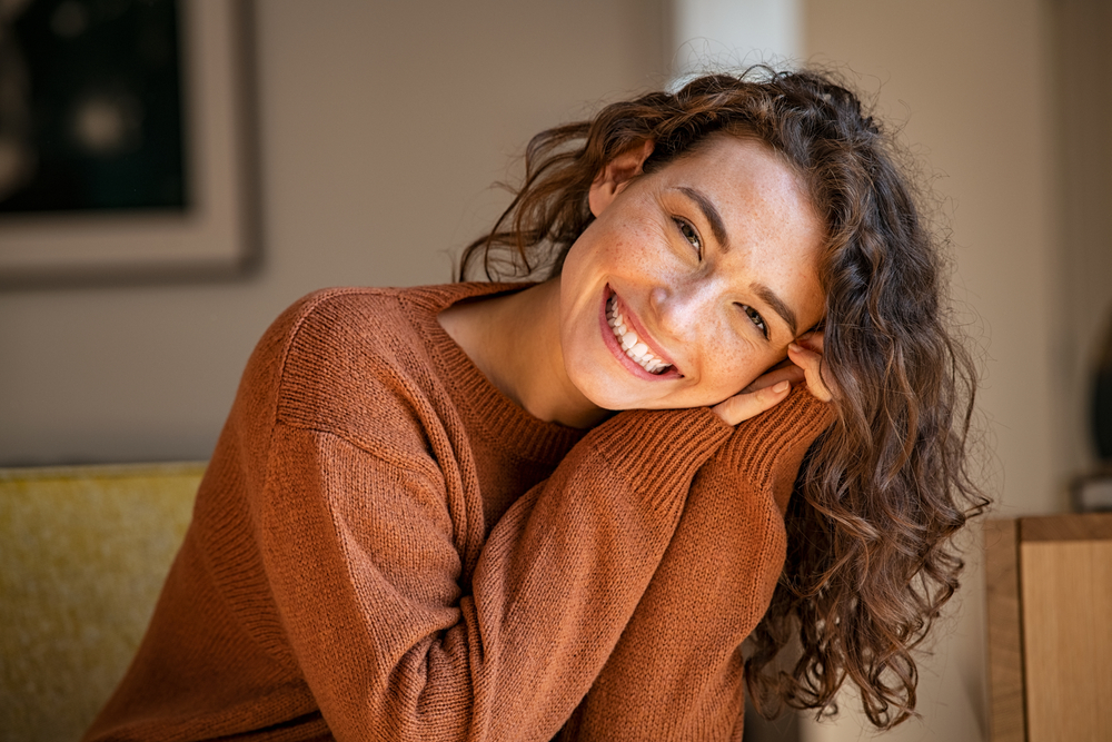 A young woman with curly brown hair smiles brightly, leaning her head on her hands. She wears a rust-colored sweater and sits indoors in soft natural light, creating a warm and cheerful atmosphere.